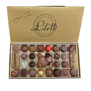 The Ultimate Holiday Sampler features an open gold box of assorted chocolates from Piletti Candies, arranged in neat rows with a variety of shapes, colors, and festive decorations.