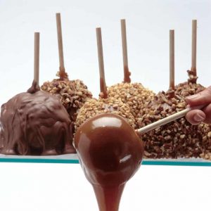 A hand dips an apple on a stick into caramel. In the background, finished caramel apples are coated with chocolate and chopped nuts.