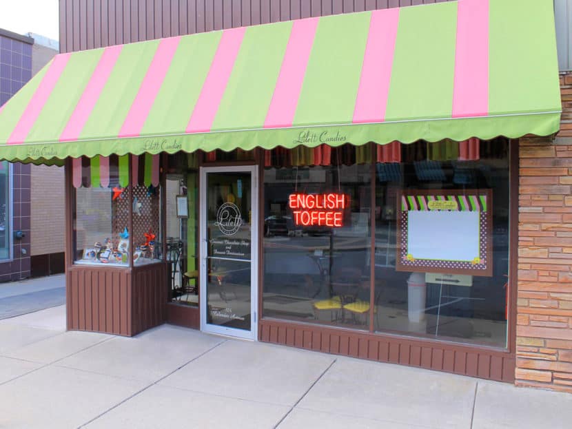 Storefront with green and pink striped awning, display window with candy, a glass door, and a neon sign reading “English Toffee.”.